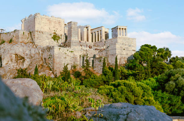 Woman wearing sunglasses, shading her face from the sun in front of the Parthenon in Greece, against blue sky.