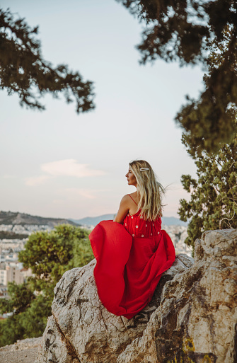 Back view of a young beautiful happy traveller female lady with a red dress and bag sitting on a rock\nand enjoys the scenery of city view landscape of cityscape view of Athens, Greece, under the brilliance of a sunny summer day, with the iconic Mount Lycabettus towering over downtown. From this vantage point, the city sprawls out beneath the hill, showcasing a blend of ancient and modern architecture. Lycabettus Hill stands as a timeless sentinel, offering a stunning backdrop to the vibrant urban landscape