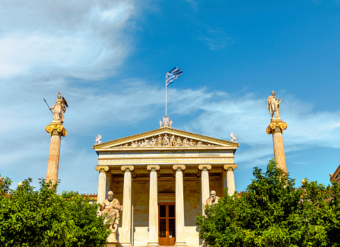 Bask in the brilliance of a sunny summer day as you behold the stately main building of the Academy of Athens, a jewel within Theophil Hansen's esteemed Trilogy, situated in central Athens, Greece. Against a backdrop of azure skies, this neoclassical masterpiece radiates timeless elegance and scholarly reverence. Its majestic columns and intricate architectural details stand in harmonious contrast to the vibrant greenery that surrounds it. As sunlight bathes the scene in a golden glow, the building invites contemplation and reflection, embodying the pursuit of knowledge and intellectual enlightenment