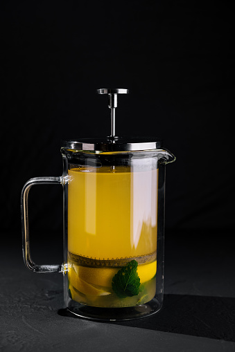 Glass french press filled with hot mint tea and lemon on a dark background