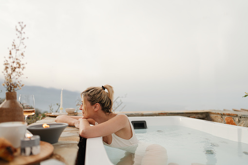 Photo of a young woman soaking in a hot tub hot tub, located on a slopes of a mountain village, overlooking the sea