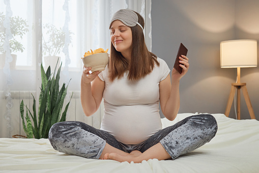 Funny smiling pregnant woman sitting in lotus pose holding bowl with potato chips and bar of chocolate smelling harmful fast food braking pregnant diet