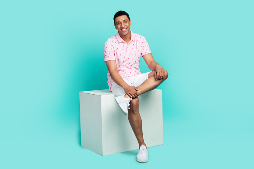 Full size photo of confident optimistic guy dressed pink polo white shorts sit on podium show shoes isolated on teal color background.