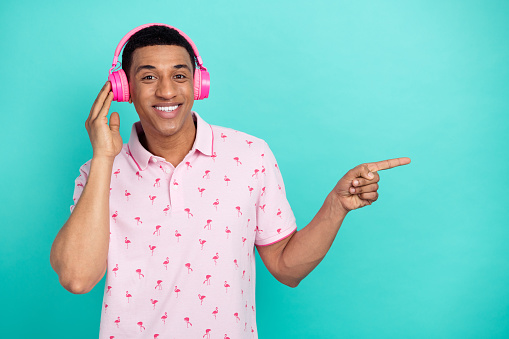 Photo of cheerful cool person toothy smile touch headphones indicate finger empty space isolated on cyan color background