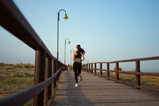 A focused young woman is running on a wooden boardwalk, her black hair streaming behind her. She is wearing athletic wear suitable for an evening workout