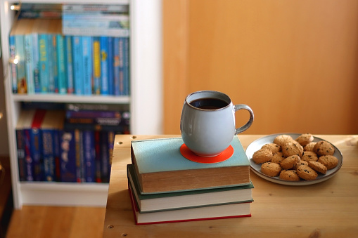 Cup of tea or coffee, pile of books, plate of cookies, reading glasses, e-reader and pen on the table. Colorful rainbow bookshelf in the background. Selective focus.