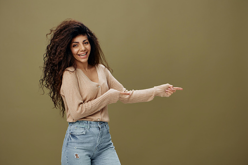 HEY LOOK HERE CONCEPT. Enjoyed smiling tanned curly Latin female in casual things point fingers aside posing isolated over olive green background. Copy space Mockup Banner. Fashion offer