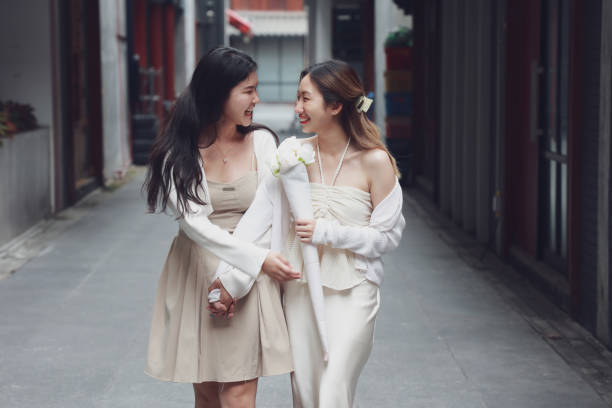 Two cheerful women walking side by side on a city street, sharing a laugh and holding flowers.