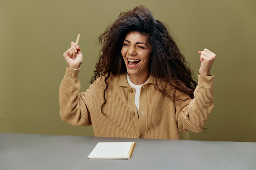 Happy overjoyed adorable tanned curly Latin lady hold hands up exult sit at the table isolated over olive green background look aside. Copy space Mockup Banner. Lucky Winner. People emotions concept