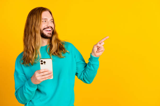 istock Portrait of funky handsome guy hold smart phone look direct finger empty space isolated on yellow color background 2148957064
