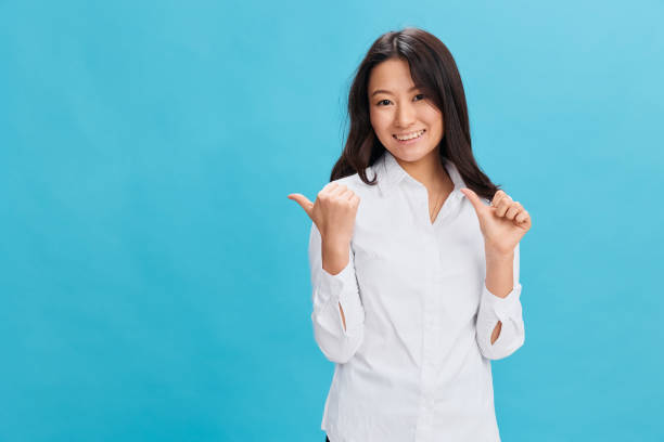 istock Smiling cute Asian businesswoman in classic office dress code point both hands aside at copy space posing isolated on over blue studio background. Cool business offer. People Emotions Business concept 2148957568
