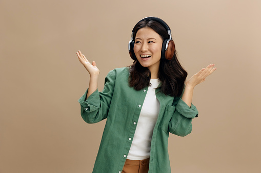 Hey look here. Cheerful happy Asian young woman in khaki green shirt headphones raise palms up posing isolated on over beige pastel studio background. Cool fashion offer. Music App Platform Ad concept