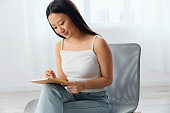 Cheerful focused young brunette writing her creation think how to make it better holds pen up looks at notebook in hand in home interior. Writer concept. Copy space Offer Banner. Full body shot