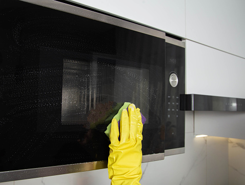 A girl's hand in a yellow glove with a sponge washes the microwave oven from grease and dirt. Foam and anti-grease.
