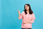 Cheerful Asian student young lady in pink hoodie sweatshirt wear round glasses point fingers aside posing isolated on over blue background. The best offer for ad. Eyewear for vision correction concept