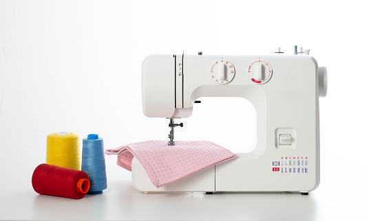 Modern automatic white sewing machine with pink fabric and multi-colored spools of thread on the table. White background. Tailoring and clothing repair services.