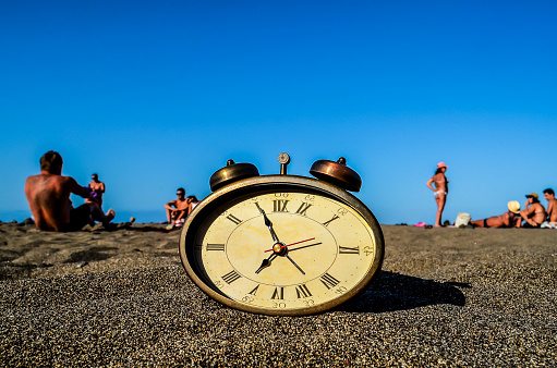 A clock is sitting on the sand with the hands on the number 12 and 3. The beach is full of people enjoying the day