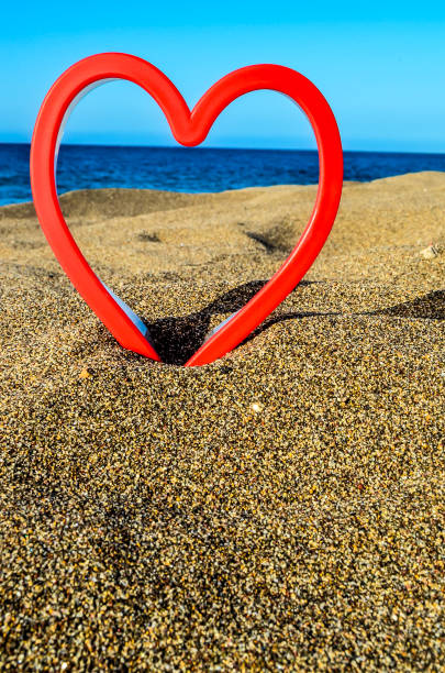 A red heart is placed on the sand at the beach. The heart is surrounded by the sand, and the beach is a beautiful blue color. Concept of love and warmth, as the heart is a symbol of affection