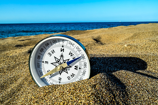 A compass is sitting on the sand, with the blue needle pointing to the north. The compass is surrounded by the ocean, creating a serene and peaceful atmosphere
