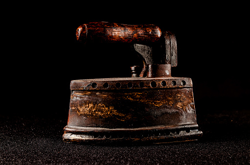 An old fashioned iron with a wooden handle sits on a black background. The iron is rusted and worn, giving it a vintage and nostalgic feel