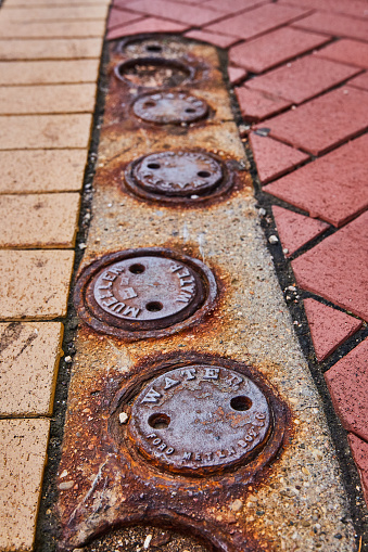 Rustic water utility covers in a row on brick pavement, symbolizing urban decay and infrastructure in Downtown Muncie, Indiana, 2023
