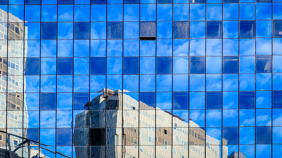 Facade of a skyscraper in which the surrounding buildings and clouds and blue sky are reflected.