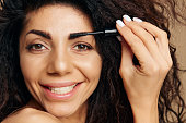 VISAGE BEAUTY CONCEPT. Smiling tanned pretty curly Latin lady enjoyed wear mascara eyebrow gel finish makeup for party posing isolated on pastel beige background, look at camera. Copy space banner