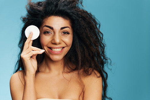 Pretty curly african american lady holding cotton pad for face, cleansing face with toner after shower over blue studio background. Happy woman enjoying fresh moisturized face skin. Copy space