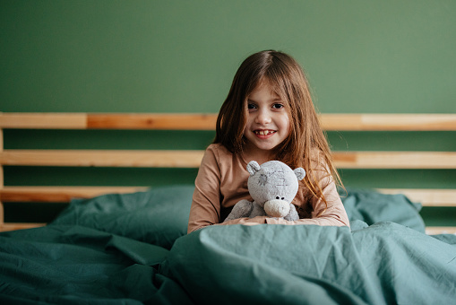 Portrait of a cute little smiling girl sitting in bed under the covers, holding a plush teddy bear and looking at the camera.