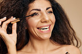 NATURAL MAKEUP CONCEPT. Joyful tanned pretty curly Latin lady wear mascara hold brush near eyelashes getting ready for party posing isolated on pastel beige background, smiling aside. Closeup banner