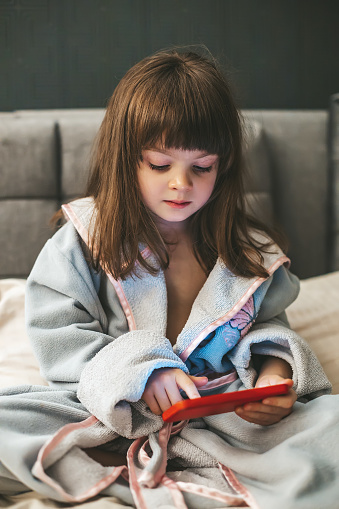 A little girl sitting on the bed, fixated on her smartphone as she interacting with digital content. Concept: kids and smartphones