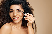 VISAGE BEAUTY CONCEPT. Smiling tanned pretty curly Latin lady enjoyed wear mascara eyebrow gel finish makeup for party posing isolated on pastel beige background, look at camera. Copy space banner