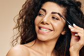 NATURAL MAKEUP CONCEPT. Smiling happy beautiful tanned curly Latin lady enjoyed wear eyebrow gel finish makeup posing isolated on pastel beige background, look aside. Copy space banner cosmetic offer