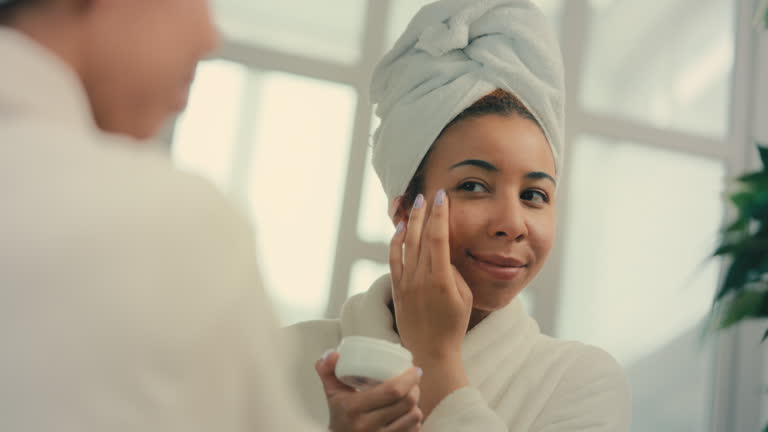 Happy woman in head towel applying undereye cream in front of mirror, skincare
