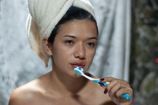 A young woman with a towel-wrapped head maintains dental hygiene, brushing her teeth in a mirror-clad, misty bathroom.