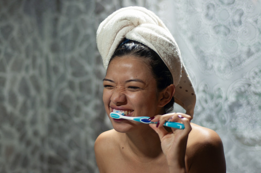 A woman, towel-wrapped hair, cringes as she diligently brushes her teeth.