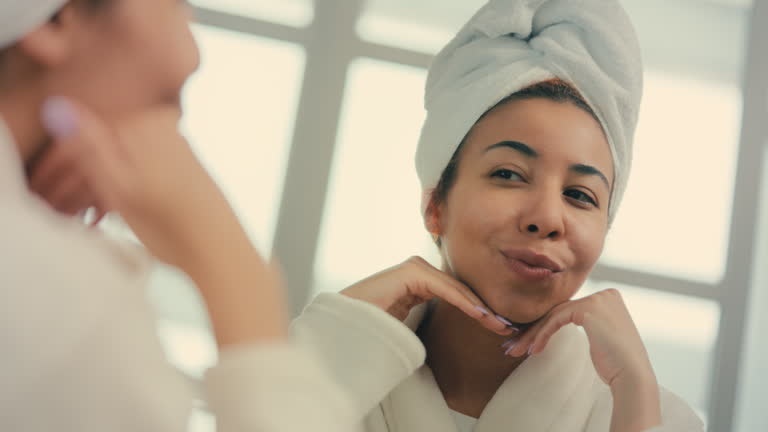 Confident young woman in head towel applying moisturizing face cream, skincare