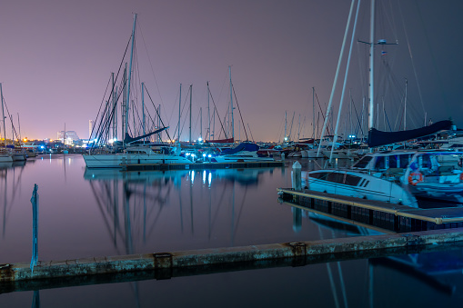 Night view of a boat dock with calm water and many boats and distant city lights in background