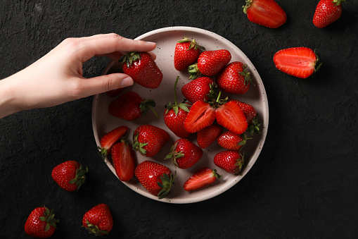 Strawberries on plate and in female hand on black background, top view