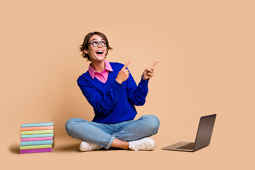 Full size portrait of astonished girl sit floor laptop book look indicate fingers empty space ad isolated on beige color background.