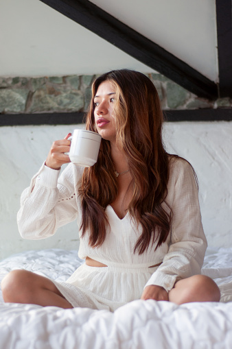 A woman enjoys a moment of tranquility, sipping warm coffee while seated comfortably on a plush bed, bathed in soft morning light.