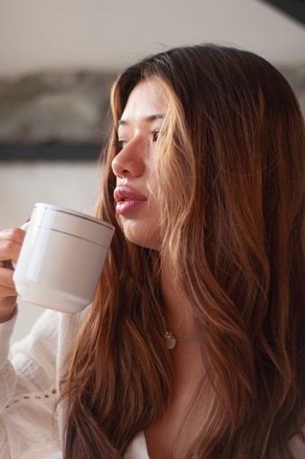 A beautiful calm woman enjoys a cup of hot coffee, embracing a peaceful moment of relaxation and self-care with a gentle gaze looking towards her window.