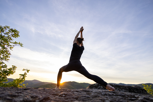 Mature woman doing yoga on top of a mountain, enjoying the sunset and the tranquility of the beautiful surroundings.