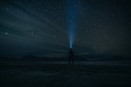 An active man wearing a headlamp looks up at the beautiful night sky filled with stars while standing on  a salt flat during an adventurous trip through Utah.