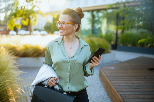 smiling modern woman worker near business center in green blouse and eyeglasses with briefcase using smartphone.