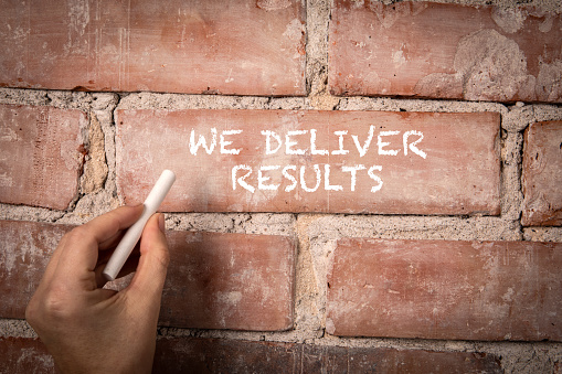 We Deliver Results. Text written with white chalk on a red brick background.