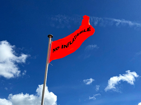 A bright orange beach warning flag blowing in the strong wind, warning (precisely because of the wind) that no inflatables should be launched; the flag is depicted against a bright blue sky.