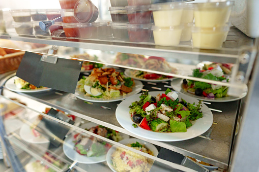 A variety of freshly prepared salads are showcased in a cafeterias food display cabinet. The salads feature vibrant greens and a mix of colorful vegetables, cheeses, and proteins, appealing to customers seeking a healthy meal option.