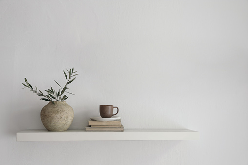 Elegant home still life. Floating shelf. Textured vase with green olive tree branches and old books. Cup of coffee, tea. Modern Mediterranean appartement. White wall background, interior indoor mockup scene.