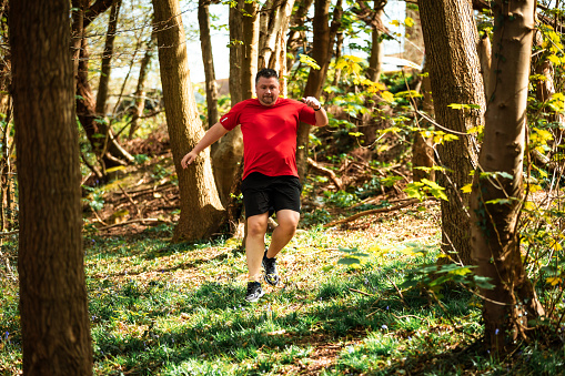 Color image depicting a man in his 30s trail running through the forest. The man is wearing sports training clothing - red t shirt, black shorts and trainers.